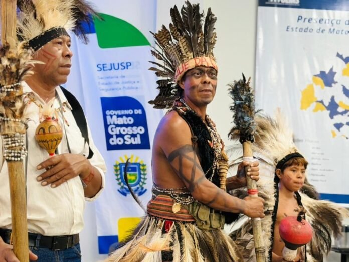 Three Indigenous Brazilians in feather headdresses and beaded outfits at a cultural event, holding ceremonial staffs.