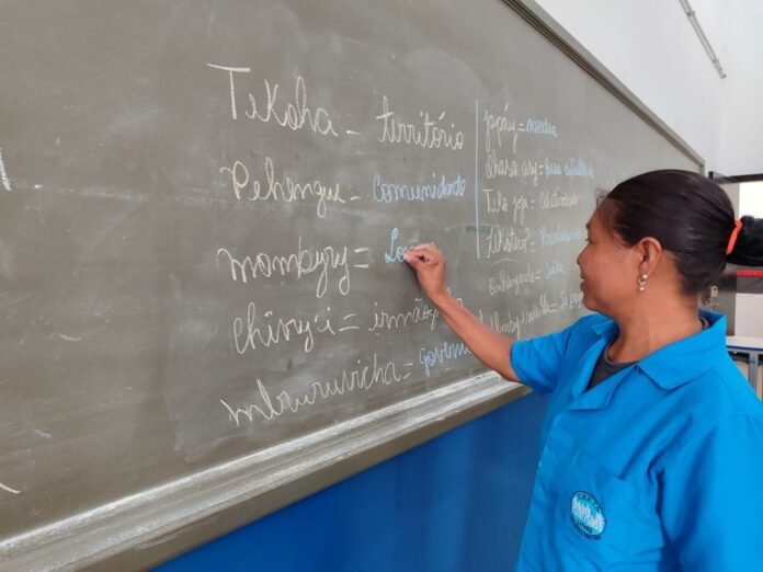 Woman in a blue uniform writing on a chalkboard in a classroom, using chalk to list words.