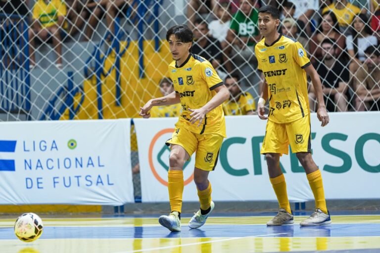 Two futsal players in yellow chase the ball on an indoor court, with a cheering crowd behind a protective net.