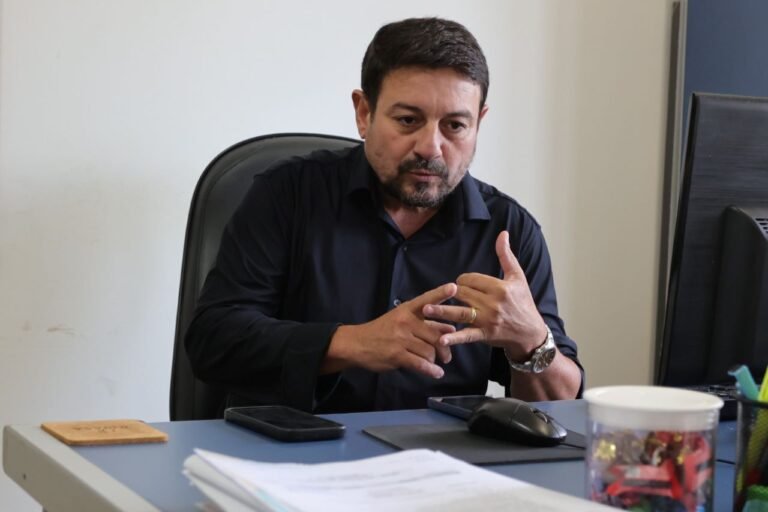 Man in a dark shirt sits at a desk, gesturing with his hands as he talks in an office setting.