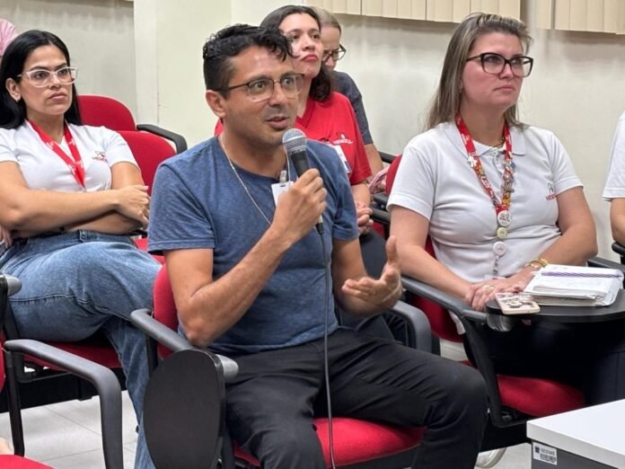 Man in blue T-shirt speaks into a handheld microphone in a panel-style meeting, with others listening and taking notes nearby.