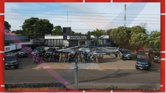 Group of police and military personnel posing in front of a Polícia Civil de Mato Grosso do Sul building, with several patrol vehicles and a helicopter nearby.