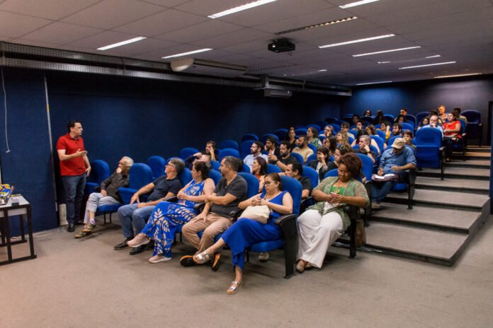 A man in a red shirt speaks to an audience seated in blue theater-style chairs in a small lecture hall.