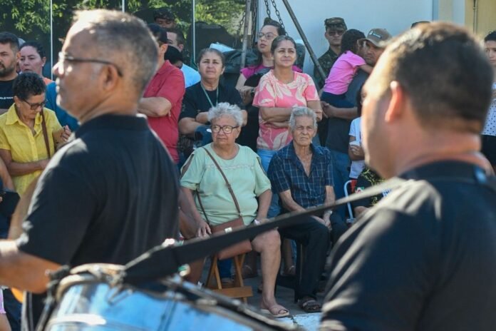 Crowd of onlookers watching an outdoor event, two elderly people seated center while others stand behind; a motorcycle in the foreground.