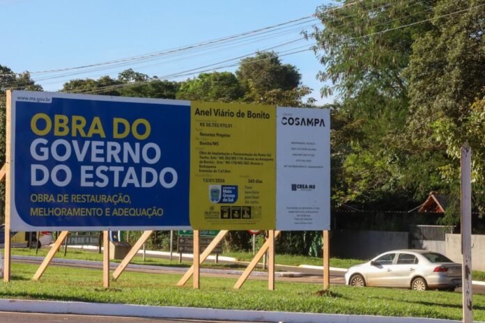 Large blue and yellow roadside billboard in Portuguese announcing state government construction work, with a white car in the background.