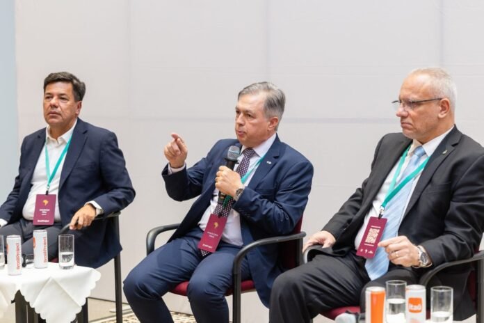 Three men in business attire sit on a panel, one holding a microphone and gesturing as others listen, credentials hanging around their necks.