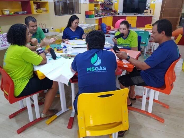 Group of adults around a round table working on crafts in a bright classroom filled with colorful shelves and toys in the background, supplies spread on the table.