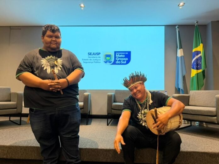 Two Indigenous men in a conference room; one stands with a carved staff, the other sits with a drum and feathered headdress, flags in the background.