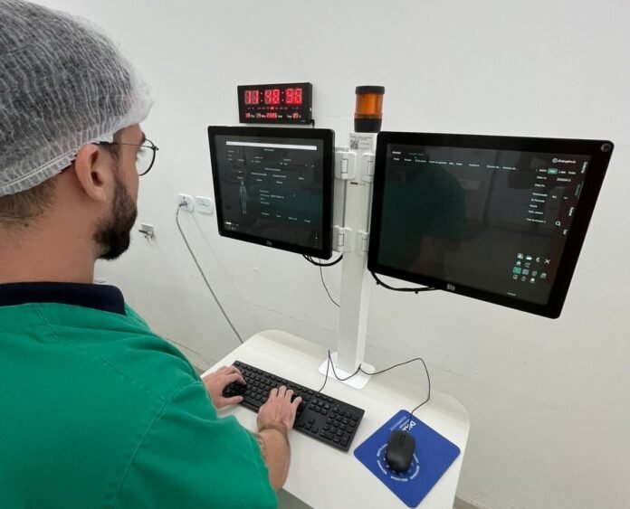 Medical professional in green scrubs and a hairnet using a dual-monitor workstation with keyboard and mouse in a clinical setting.