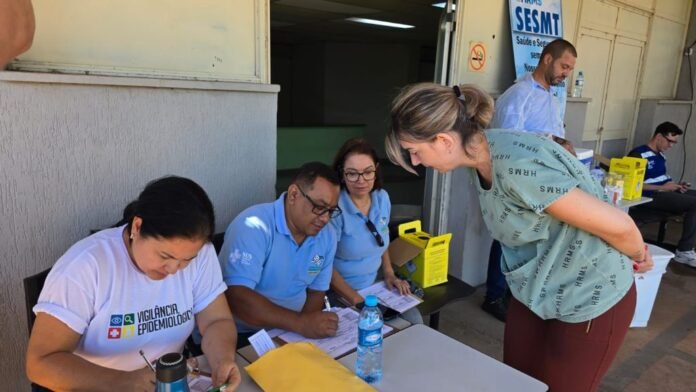 Health workers sit at a table outside a clinic, filling forms as a woman leans in to talk to them.