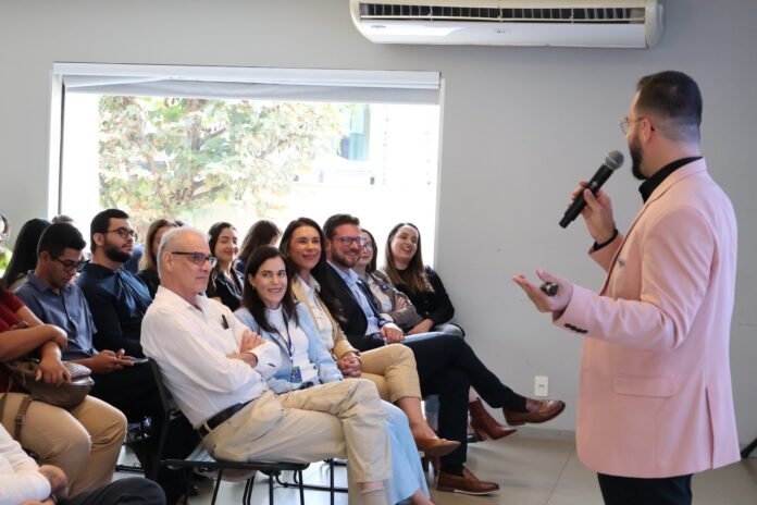 Speaker in a pink blazer addresses a seated audience in a conference room, holding a microphone and gesturing with his other hand.