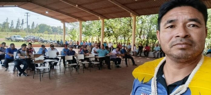 Group of people seated around desks in an open-air pavilion, with a man in a yellow shirt taking a selfie in the foreground.
