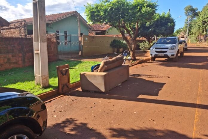 Beige sofa left on a residential street curb with a worn chair nearby and parked cars under a sunny blue sky, trees casting shadows.