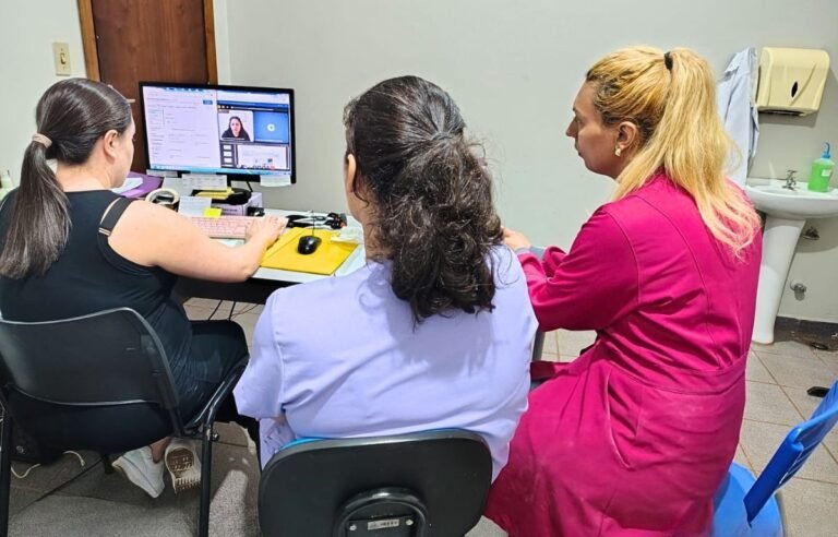 Three women sit at a desk in a clinic, looking at a computer screen showing a video call.