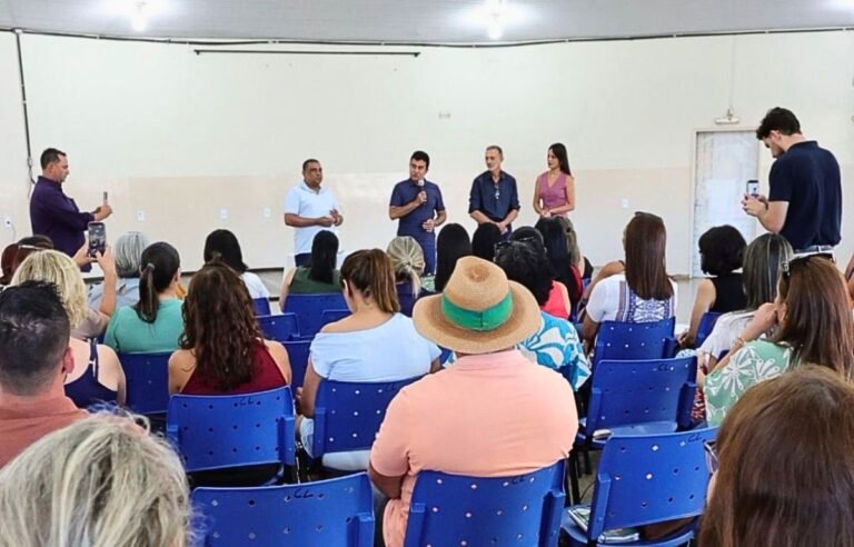 Panel of five speakers stands at the front addressing a seated audience in a bright room.