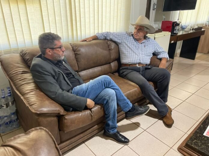 Two men sit on a brown leather couch in a lounge, chatting—one in a cowboy hat and check shirt, the other in a blazer and jeans.