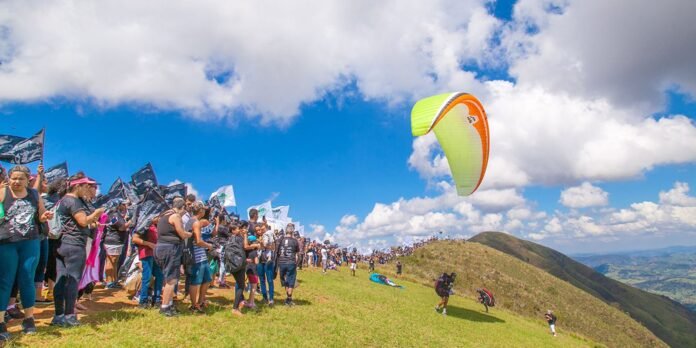 Crowd of spectators along a grassy hillside watching a paragliding event as a bright lime and orange paraglider soars in a blue sky.