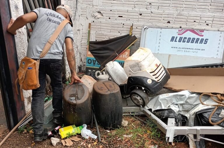 Construction worker in a gray shirt and tool belt examines rusty barrels and scattered junk outside a brick wall corner.