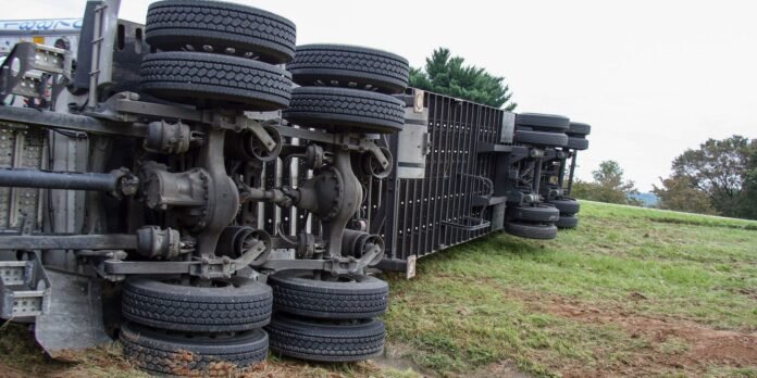 Overturned semi-trailer on its side with exposed wheels and axles, lying on a grassy slope in an open area.