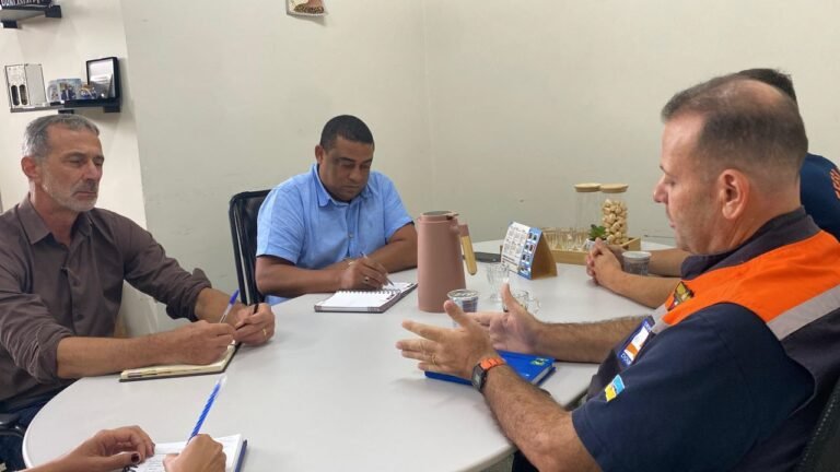 Man in an orange safety vest speaks with open hands as three colleagues take notes during a meeting at a white table in a small office setting with a pink pitcher nearby. (informative)