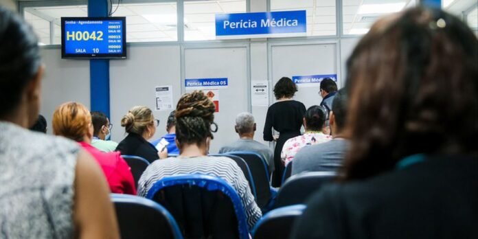 People seated in a medical clinic waiting area facing a receptionist/exam area with a blue sign reading 'Perícia Médica'.