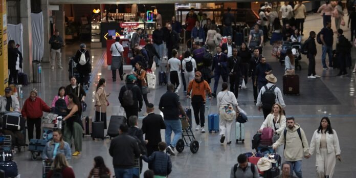 Crowded airport-like terminal with travelers pushing suitcases, backpacks, and strollers along a shiny floor and shopfronts in the background.