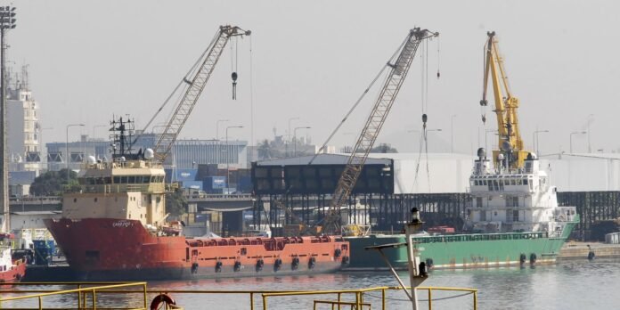 Port scene with a red-hulled cargo vessel on the left and a green-hulled vessel on the right, docked beside tall cranes and industrial structures.