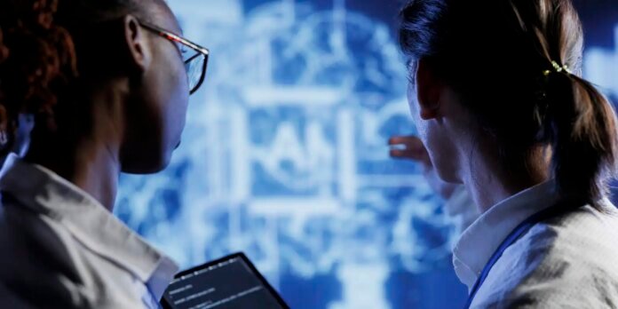 Two researchers in lab coats study a large blue digital display with schematic diagrams, viewing from behind.