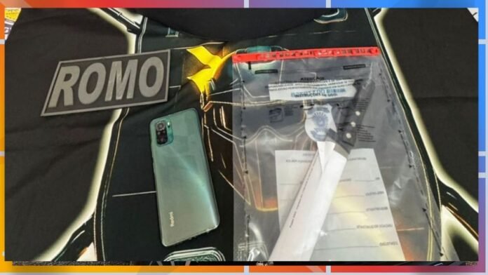 Green smartphone resting on a dark surface beside a clear zippered document pouch with papers, with a 'ROMO' patch/logo visible in the background.