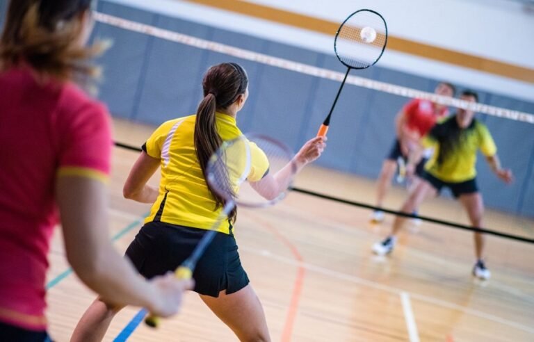Woman in a yellow shirt swings a badminton racket to hit the shuttlecock on an indoor court; players in the background.
