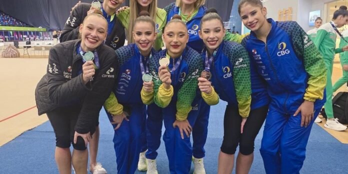 Group of female gymnasts posing with medals after a competition, wearing blue and green team jackets on a gym floor.