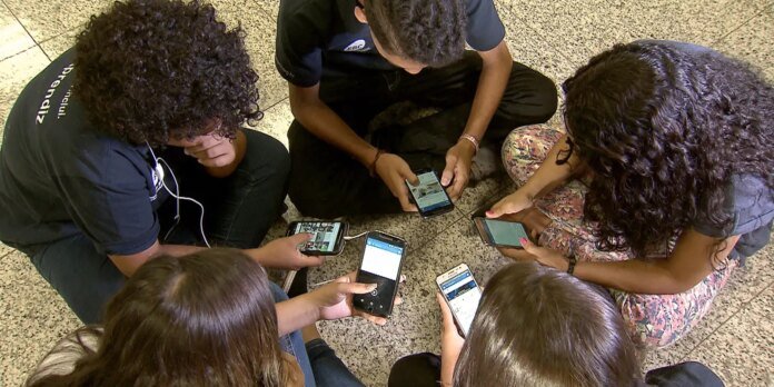 Group of six people sitting in a circle on a speckled tile floor, each person looking at a smartphone.