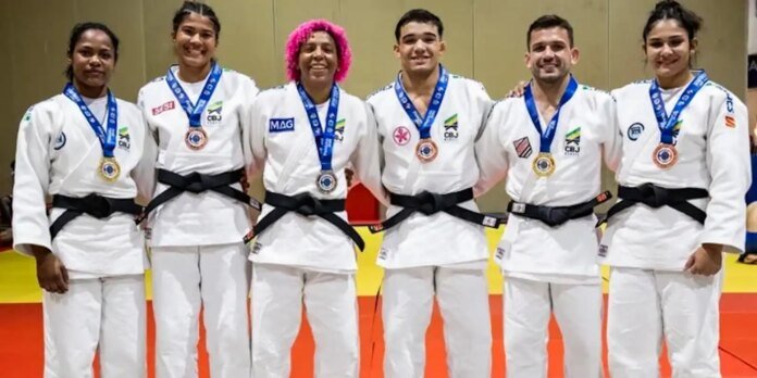 Six judokas in white uniforms with black belts pose together on a competition mat, each wearing a medal around the necks after a event.