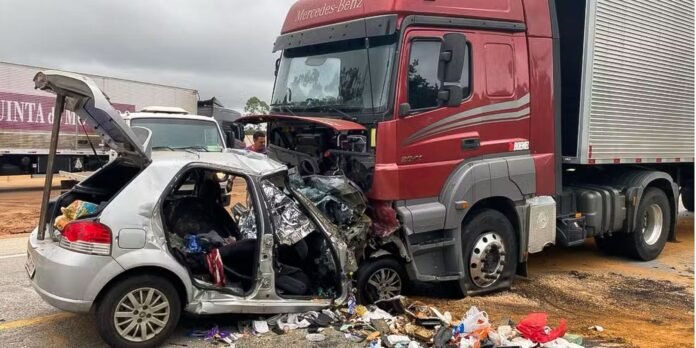 Silver hatchback heavily crushed at the front of a red semi-truck after a collision, debris scattered on the road nearby.