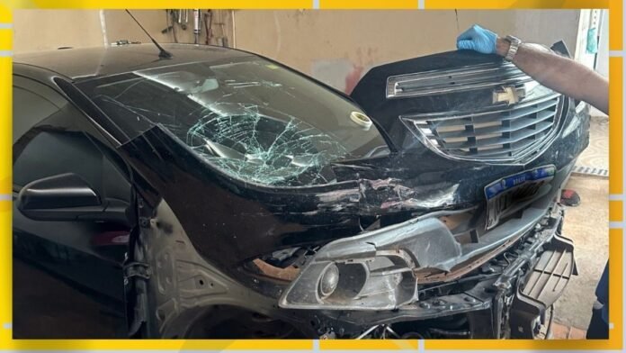 Black SUV with severe front-end damage, shattered windshield, and crumpled hood in a workshop; a gloved hand touches the damaged grille area.