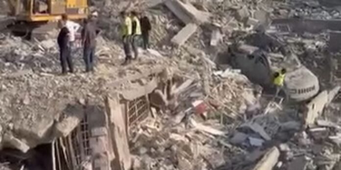 Rubble and debris from a collapsed building with construction machinery in the background and a few workers in safety vests standing on the site.