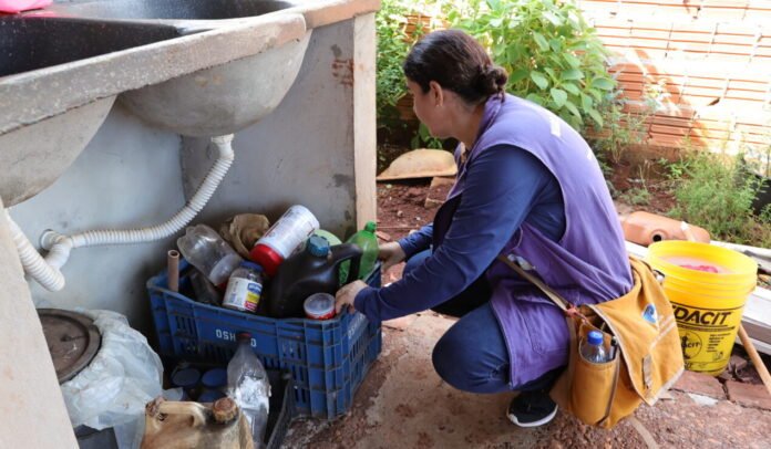 Woman kneeling beside a blue crate filled with plastic bottles and containers under an outdoor sink, sorting recyclables.