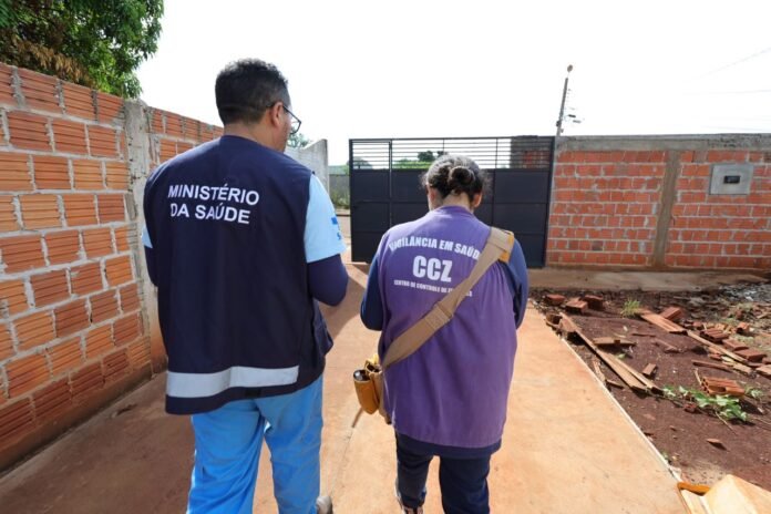 Two health workers walk away from the camera along a brick-walled path; one wears a navy vest that reads 'MINISTÉRIO DA SAÚDE' and blue pants, the other in a purple shirt with 'VIGILÂNCIA EM SAÚDE CCZ'.