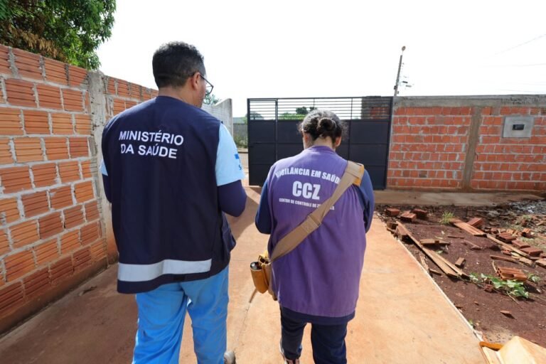 Two health workers walk away from the camera along a brick-walled path; one wears a navy vest that reads 'MINISTÉRIO DA SAÚDE' and blue pants, the other in a purple shirt with 'VIGILÂNCIA EM SAÚDE CCZ'.