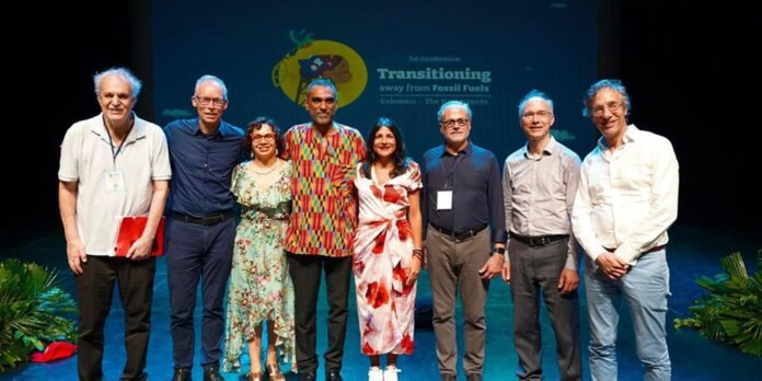 Eight diverse people stand together on a stage for a conference photo, with a banner reading Transitioning away from Fossil Fuels in the background.