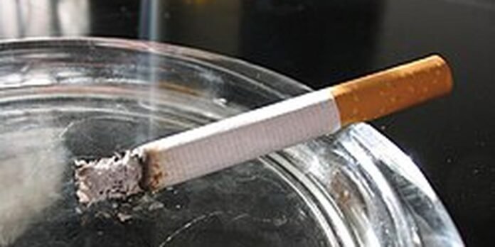 Cigarette butt resting on the rim of a glass ashtray, with ash nearby.