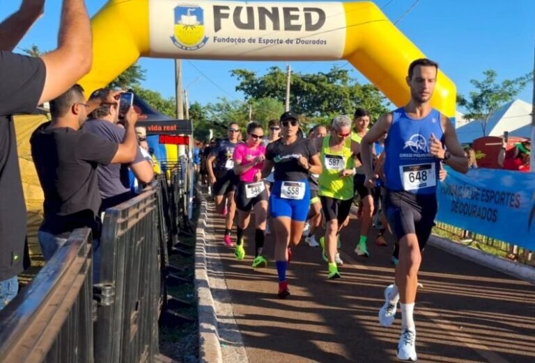 Runners start a road race under a yellow inflatable arch with FUNED text; bib numbers visible, spectators on the left taking photos.