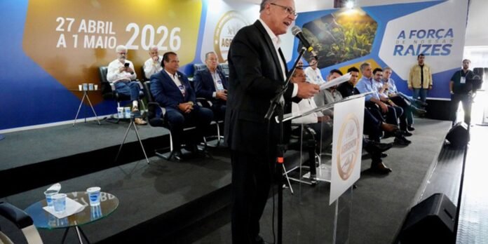 Man in a dark suit speaks at a podium on a stage during a conference; backdrop shows the date '27 ABRIL A 1 MAIO 2026' and the slogan 'A FORÇA DE NOSSAS RAIZES'. Behind him, seated attendees watch.