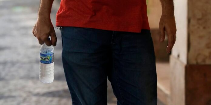 Person stands outdoors wearing a red shirt and dark jeans, holding a plastic water bottle in their left hand.
