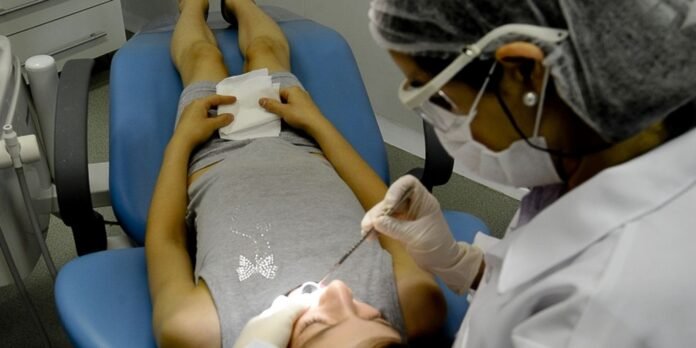 Dentist wearing a mask and gloves treats a patient lying in a dental chair, using a tool near the mouth.