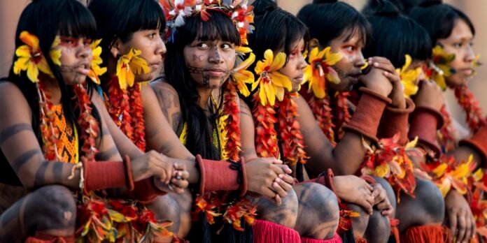 Group of women in traditional dress with colorful flower headpieces and body paint, seated arm-in-arm in a line at a cultural ceremony.],