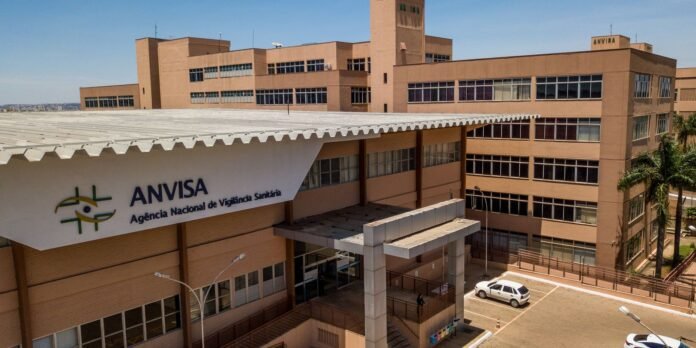 Exterior view of ANVISA headquarters (Agência Nacional de Vigilância Sanitária) — beige brick building with large windows, entrance canopy, and parked cars nearby.
