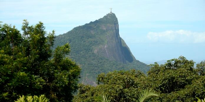 Corcovado mountain with Christ the Redeemer statue at the summit, surrounded by lush forest under a blue sky.