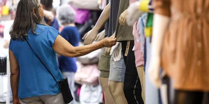 Older woman in a blue blouse reaches to touch a gray skirt on a clothing rack in a busy store.