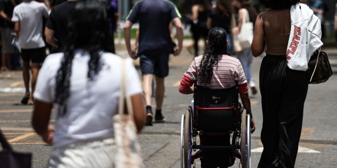 A person in a wheelchair seen from behind, rolling through a crowded city street with others walking nearby.